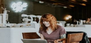 Young woman with long red hair, wearing a brown v neck, short sleeved top, is sat down working on her laptop.